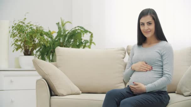 4k video av gravida kvinna sitter på vit soffa och visar tummen upp. — Stockvideo
