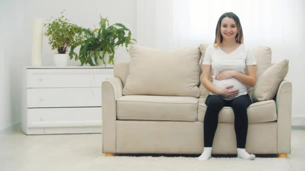 4k-Video von schwangerer Frau, die auf weißem Sofa sitzt und ihren Bauch berührt. — Stockvideo