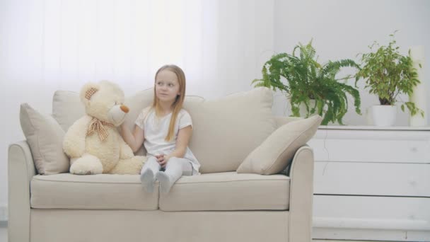 Vidéo ralenti 4k de petite fille partageant des secrets avec un ours en peluche sur le canapé. — Video