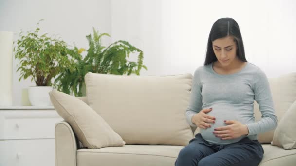 4k video van zwangere vrouw zitten op witte bank en het aanraken van haar maag. — Stockvideo