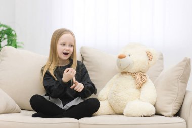 Küçük kız beyaz oyuncak ayısıyla kanepede sohbet ediyor..