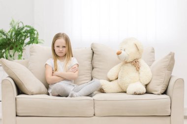 Photo of sad little blond girl in white clothes near teddy bear. clipart