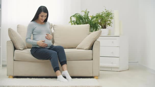 4k slow motion video av gravida kvinna som sitter på vit soffa och vidrör magen. — Stockvideo