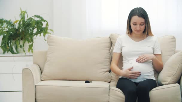 4k video de la mujer embarazada sentada en un sofá blanco y tocando su estómago. — Vídeo de stock