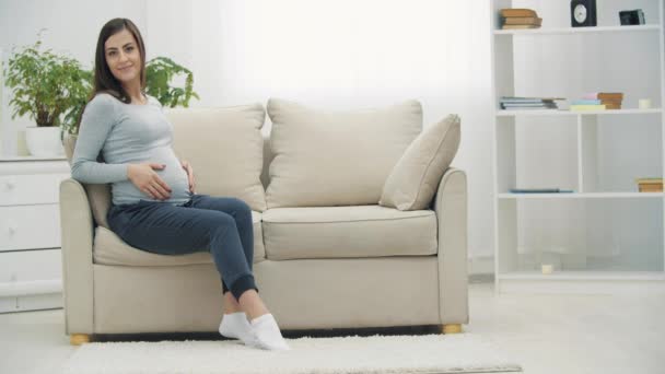 4kスローモーションビデオの妊娠中の女性座っています上の白いソファと表示親指. — ストック動画