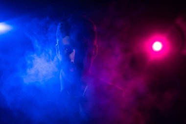 Fotoğraf: Pembe ve mavi ışıklarla arka planda kameraya bakan adam..