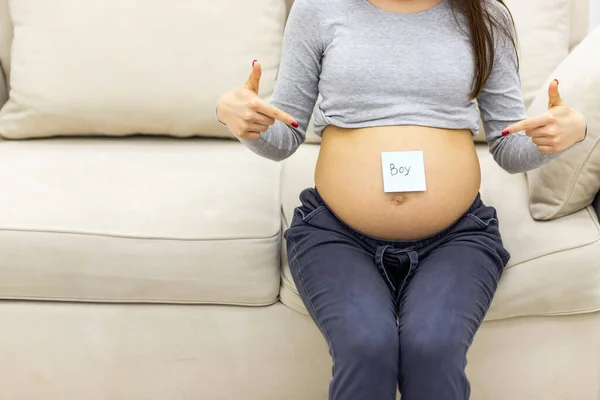 Foto recortada de la mujer embarazada sentada en el sofá con un papel en el estómago. —  Fotos de Stock