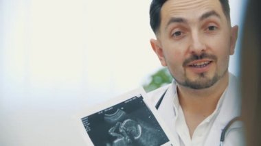 Doktorun beyaz laboratuvar önlüğü giymiş, elinde ultrason fotoğrafları olan ve açıklama yapan 4K yavaş çekim videosu. Doktorluk mesleği kavramı.