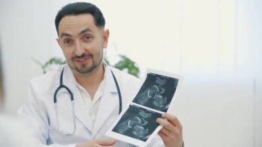 Doktorun beyaz önlük giyip ultrason fotoğraflarını tutarken ve açıklama yaparken 4K videosu. Doktorluk mesleği kavramı.