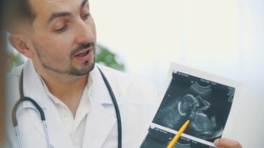 Doktorun beyaz önlük giyip ultrason fotoğraflarını tutarken ve açıklama yaparken 4K videosu. Doktorluk mesleği kavramı.