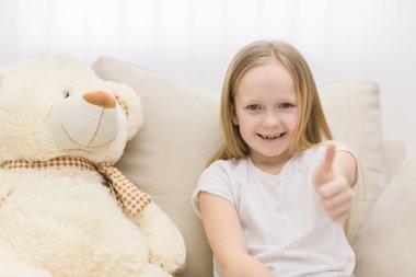 Photo of teddy bear and little girl showing thumb up at the camera. Concept of cute little girl.