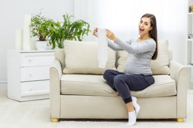 Hamile bir kadının kucağında küçük kıyafetlerle fotoğrafı. Hamile kadın kavramı.