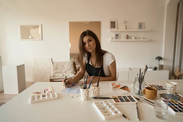 Linda artista feminina em seu estúdio. Desenho feminino em álbum com tintas aquarela e pincel. — Fotografia de Stock