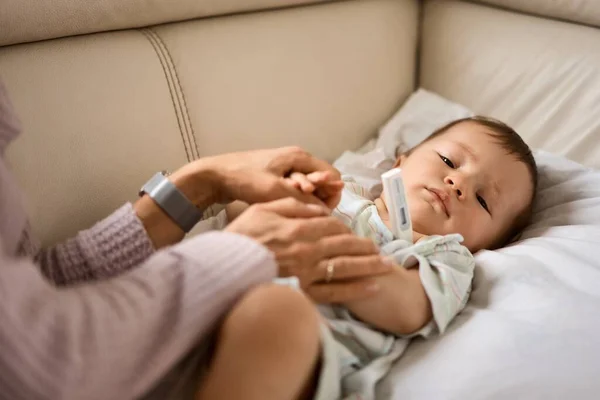 Ibu muda yang khawatir duduk di sofa disamping anaknya yang sakit demam tinggi. Ibu mengukur suhu menggunakan termometer anak sakit yang terbaring di bawah selimut di rumah. — Stok Foto