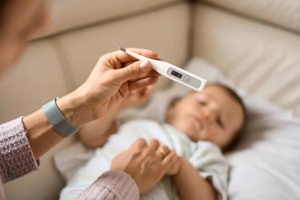 Ibu muda yang khawatir duduk di sofa disamping anaknya yang sakit demam tinggi. Ibu mengukur suhu menggunakan termometer anak sakit yang terbaring di bawah selimut di rumah. — Stok Foto