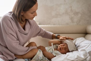 Onun baş ve ölçüm sıcaklığı termometre ile üzerinde soğutucu jel ile genç küçük çocuk kanepede. ve kendine iyi bak onu anne tarafından