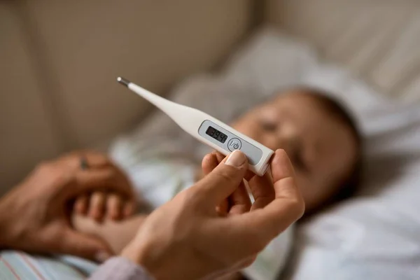 Endişeli genç anne, hasta oğlunun yanında, yüksek ateşi olan kanepede oturuyor. Annem evde battaniyenin altında yatan hasta çocuğun termometresini kullanarak sıcaklık ölçer.