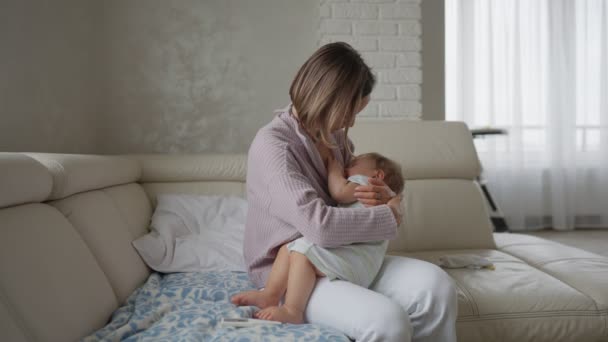 Worried young mother sitting on sofa beside her sick son with high fever. Mom measures temperature using thermometer of sick child lying under blanket at home — Stock Video