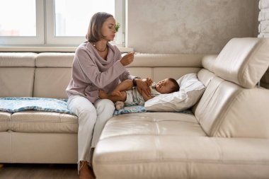 Endişeli genç anne, hasta oğlunun yanında, yüksek ateşi olan kanepede oturuyor. Annem evde battaniyenin altında yatan hasta çocuğun termometresini kullanarak sıcaklık ölçer.