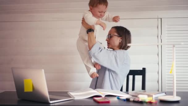 Genç bir anne bebeğiyle ilgilenirken evde bilgisayar kullanarak çalışıyor. Kilitleme sırasında evden çalış — Stok video
