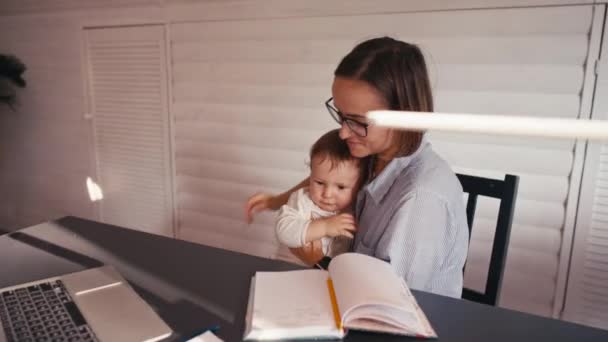 Молодая мать работает дома за компьютером, в то время как она заботится о своем ребенке. Работа из дома во время изоляции — стоковое видео