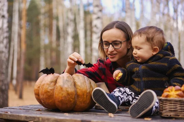 Молодая женщина и ее маленький сын в осеннем парке, мальчик играет с тыквой Хеллоуина и ест тыквенную булочку. — стоковое фото