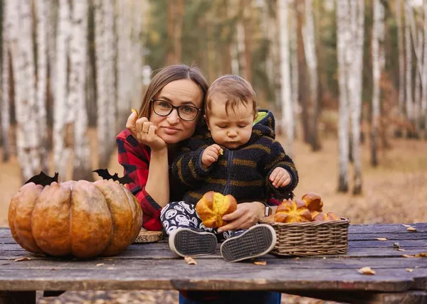 Молодая женщина и ее маленький сын в осеннем парке, мальчик играет с тыквой Хеллоуина и ест тыквенную булочку. — стоковое фото