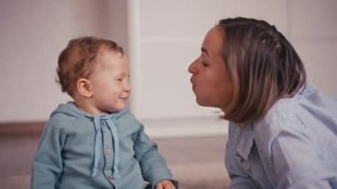 Anne ve küçük oğlu evde eğleniyor ve oynuyorlar. 1 yaşındaki küçük bir çocuk evde annesiyle oynuyor ve yerde oturuyor. Mutlular, gülümsüyorlar ve gülüyorlar..