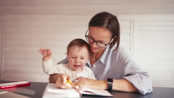 Genç bir anne bebeğiyle ilgilenirken evde bilgisayar kullanarak çalışıyor. Kilitleme sırasında evden çalış — Stok video