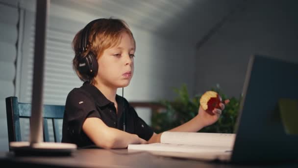 Дистанційне навчання, хлопчик в навушниках сидить за столом вдома, дивлячись на буковий ноутбук. Концепція онлайн-освіти, додому. Маленький хлопчик робить домашнє завдання вдома пізно ввечері . — стокове відео