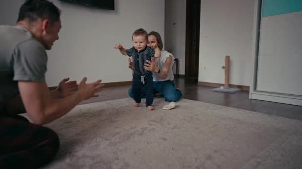 Bonne famille souriant comme bébé fait ses premiers pas. La famille à la maison encourage le bébé fils à prendre les premières mesures — Video