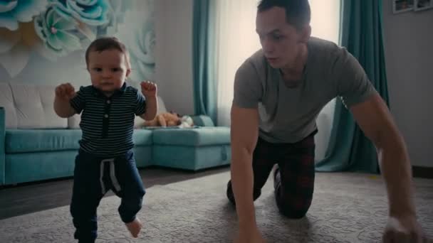 Baby macht erste Schritte mit Vätern Hilfe zu Hause. Vater ermutigt kleinen Sohn zu ersten Schritten zu Hause — Stockvideo