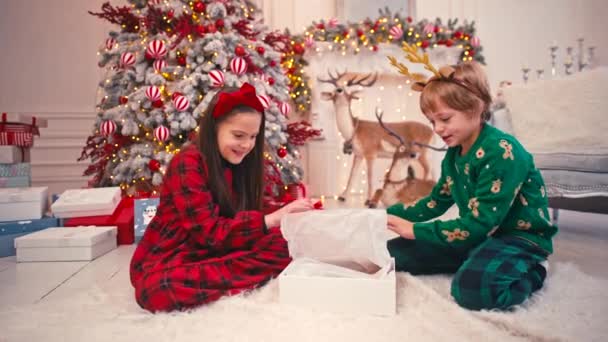 Schöne Kinder, die in der Nähe des Weihnachtsbaums spielen und Schachteln mit Geschenken öffnen. Wohnzimmer. Hausdekoration. Heiligabend. Familienkonzept. — Stockvideo