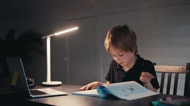 在远程学习中，一个男孩坐在桌旁，看着家里的山毛榉笔记本电脑。网上教育的概念，家。小男孩晚上很晚才在家里做作业. — 图库视频影像