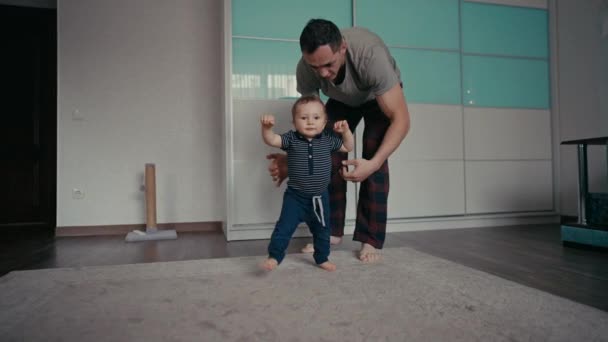 赤ちゃんは父親と一緒に最初の一歩を踏み出すと家で助けます。お父さんは赤ちゃんの息子が家で最初の一歩を踏み出すことを奨励 — ストック動画