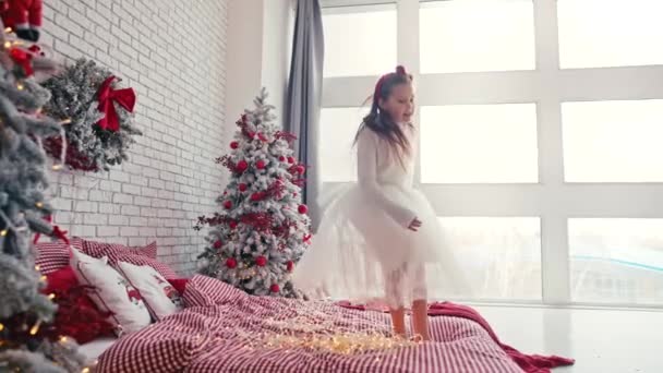 Menina bonito em vestido branco pulando na cama com travesseiro, Natal hora da manhã, festa de crianças. Natal traz tanta diversão. — Vídeo de Stock