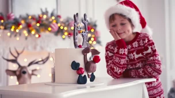 O rapaz está sentado perto do bolo de Natal. Menino à espera de Natal para comer bolo — Vídeo de Stock