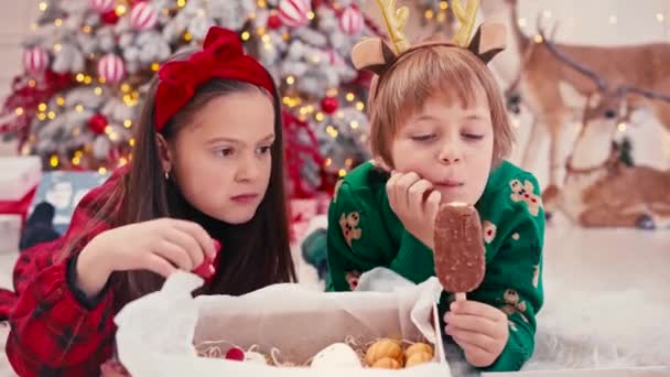 Retrato de vídeo 4k de dos niños pequeños y lindos comiendo dulces de Navidad. Dulces artesanales de chocolate hechos a mano en manos de niños. Hermano comiendo dulces cerca del árbol de Navidad — Vídeos de Stock