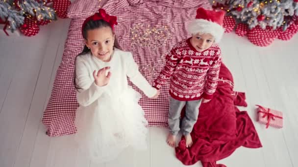 Portrait of attractive cute kids wearing christmas wear falling together on bed in apartment, laughing, raising hands and legs up. Slow motion shot — Stock Video