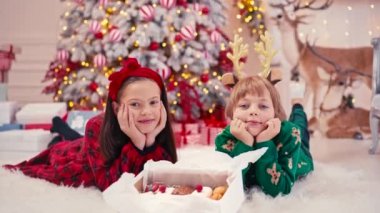 Noel şekeri yiyen iki küçük çocuğun 4K video portresi. El yapımı çikolatalı tatlılar çocukların elinde. Kardeşim Noel ağacının yanında şeker yiyor.
