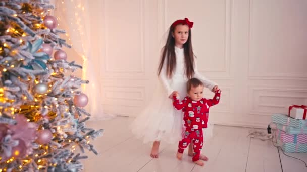 Ребенок делает первые шаги дома, держась за руки с сестрой в белом платье. — стоковое видео