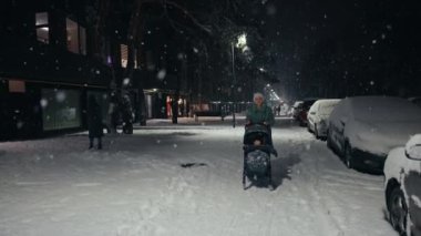Young mother pushing baby stroller and walking at city street during snowfall. Snow covered trees and bushes. Spending time with baby in beautiful winter night. Enjoying peaceful stroll