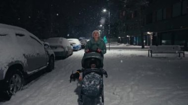 Young mother pushing baby stroller and walking at city street during snowfall. Snow covered trees and bushes. Spending time with baby in beautiful winter night. Enjoying peaceful stroll