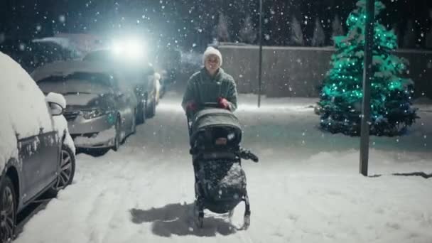 Young mother pushing baby stroller and walking at city street during snowfall. Snow covered trees and bushes. Spending time with baby in beautiful winter night. Enjoying peaceful stroll — Videoclip de stoc