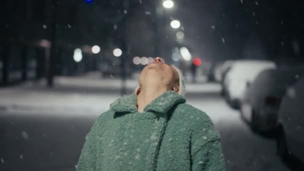 Cinematic shot of carefree happy smiling young woman wearing warm hat is excited and amazed with snowfall in a festive winter evening.Concept of winter holidays, freedom, happiness. — Stockvideo