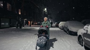Young mother pushing baby stroller and walking at city street during snowfall. Snow covered trees and bushes. Spending time with baby in beautiful winter night. Enjoying peaceful stroll