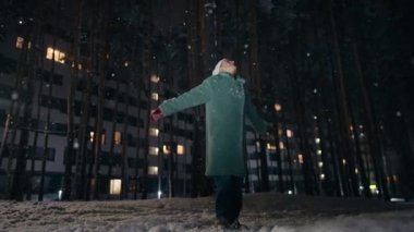 Cinematic shot of carefree happy smiling young woman wearing warm hat is excited and amazed with snowfall in a festive winter evening.Concept of winter holidays, freedom, happiness.