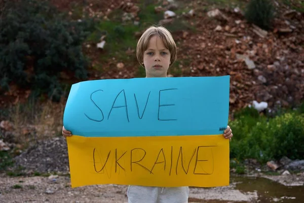 Retrato de niño ucraniano cerca del edificio destruido mostrando pancarta con texto de masaje Save Ukraine. Pidiendo ayuda. Crisis, guerra, no paz, detener la agresión de Rusia país —  Fotos de Stock