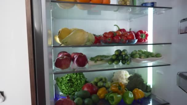 冷蔵庫から生食を取っている女性。健康的な食べ物でいっぱいの冷蔵庫。果物や野菜. — ストック動画