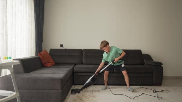 Девятилетний мальчик веселится во время уборки. Мальчик держит пылесос, как гитару, и танцует с ним, притворяясь, что играет, слушая музыку в наушниках — стоковое видео